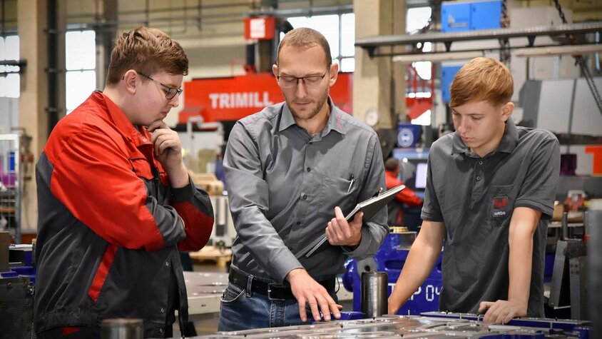 Das Bild zeigt den Lehrlingsausbilder, der zwei Lehrlingen in der Werkzeugbautechnik in einer Produktionsumgebung ein Stanz- und Umformwerkzeug erklärt. Er zeigt auf bestimmte Teile des Werkzeugs und erklärt die Funktionen und den Zweck jeder Komponente. Die Lehrlinge hören aufmerksam zu und stellen Fragen, während sie lernen. Die Szene vermittelt den Eindruck von Wissensaustausch und praktischer Schulung in einem professionellen Arbeitsumfeld.