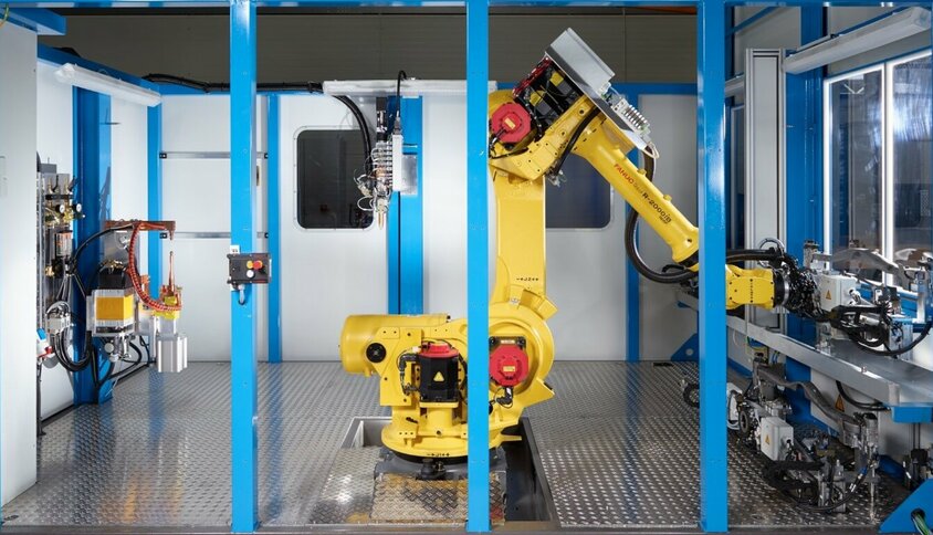 ine moderne Fügetechnik-Anlage mit einem gelben FANUC-Roboterarm in einem abgeschirmten Arbeitsbereich. Der Roboter ist zentral in der Anlage positioniert und bereit für automatisierte Klebeaufgaben. Umgeben von blauen Rahmenstrukturen zeigt die Szene eine hochmoderne Fertigungsumgebung mit präzisen mechanischen Komponenten und fortschrittlichen Steuergeräten. Diese Anlage steht für Präzision, Effizienz und Automation in der Fügetechnik.