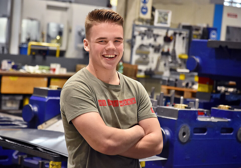 Ein Lehrling bei weba Werkzeugbau, fotografiert in der Produktionshalle, lächelt direkt in die Kamera. Er trägt ein T-Shirt mit der Aufschrift 'We are WEBA', was seinen Stolz und seine Zugehörigkeit zum Unternehmen zum Ausdruck bringt. Im Hintergrund sind mehrere Werkbänke und Teile eines dunkelblauen Umformwerkzeugs sichtbar, was auf die praktische und lernintensive Umgebung hinweist, in der der Lehrling ausgebildet wird. Das Bild strahlt Jugendlichkeit, Optimismus und das Engagement des Lehrlings für seine Ausbildung aus, während es gleichzeitig die unterstützende und lehrreiche Atmosphäre bei weba hervorhebt. Das Bild steht symbolisch für Lehrstellen Jobs in Steyr bei weba Werkzeugbau.