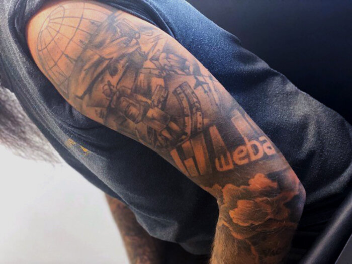 A subtly colored upper arm tattoo of an employee featuring the logo of the company "weba." Surrounding the logo are detailed technical elements like gears, machines, and a globe. The tattoo symbolizes the employee's strong connection to weba Werkzeugbau and their passion for technology and precision.