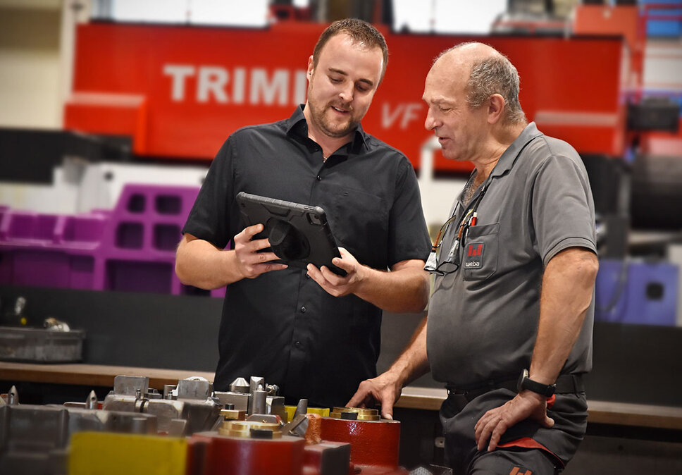 Two weba employees during an on-site technical consultation. They are standing at a workbench with tool parts, discussing details using a tablet. The mobile toolmaker service ensures expert consultation and optimal adjustment of the tools to the production processes.