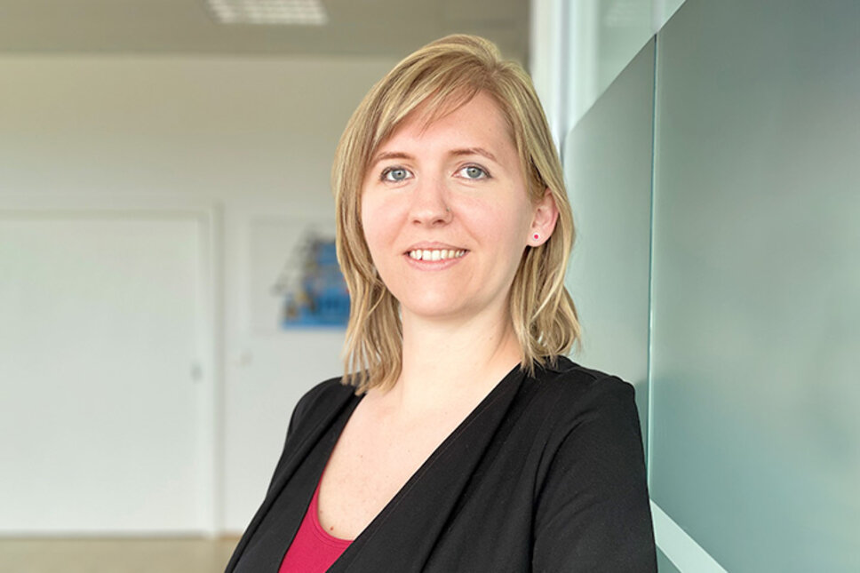 Portrait of Tanja Burghuber, Human Resources Manager at weba Werkzeugbau, leaning relaxed against a frosted glass wall in Dietach, with a professional and friendly expression, highlighted by soft lighting and a blurred background. The photo is used in the contact section of the job openings in Steyr at weba Werkzeugbau.