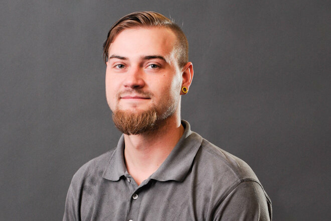 A portrait photo of Bronislav Škoda, a young man with short, side-shaved hair and a well-groomed full beard. He is wearing a gray polo shirt with the weba logo and is looking warmly into the camera. His ears are adorned with yellow plugs, and the background is a neutral gray studio backdrop.