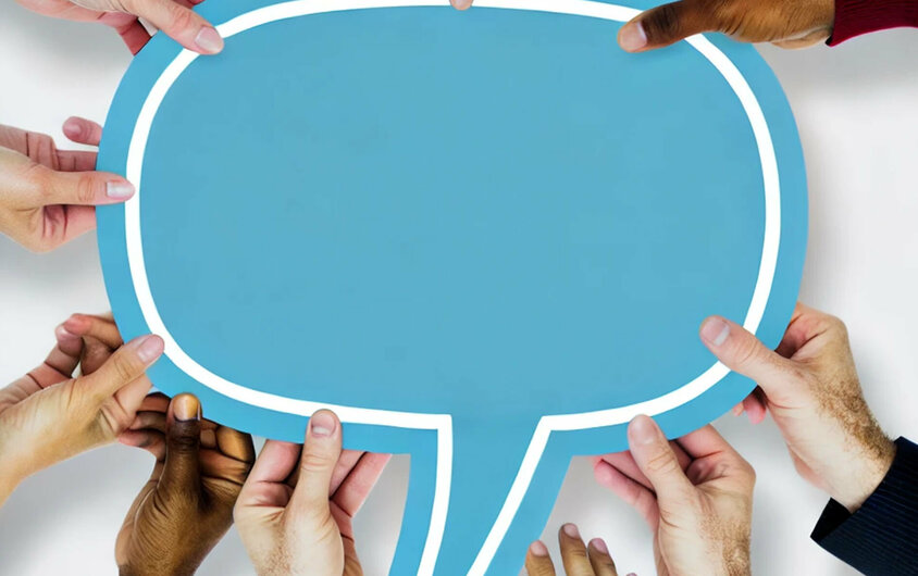 A group of diverse hands holding a large blue speech bubble together. The speech bubble is empty, symbolizing communication and collaboration. The background is plain, emphasizing the collective effort in supporting the speech bubble.