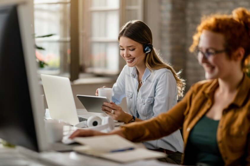 Lächelnde weibliche Person mit Headset, die Kaffee trinkt und im Büro das Touchpad nutzt. Das Bild symbolisiert glücklichen Berufseinstieg in Steyr bei weba.