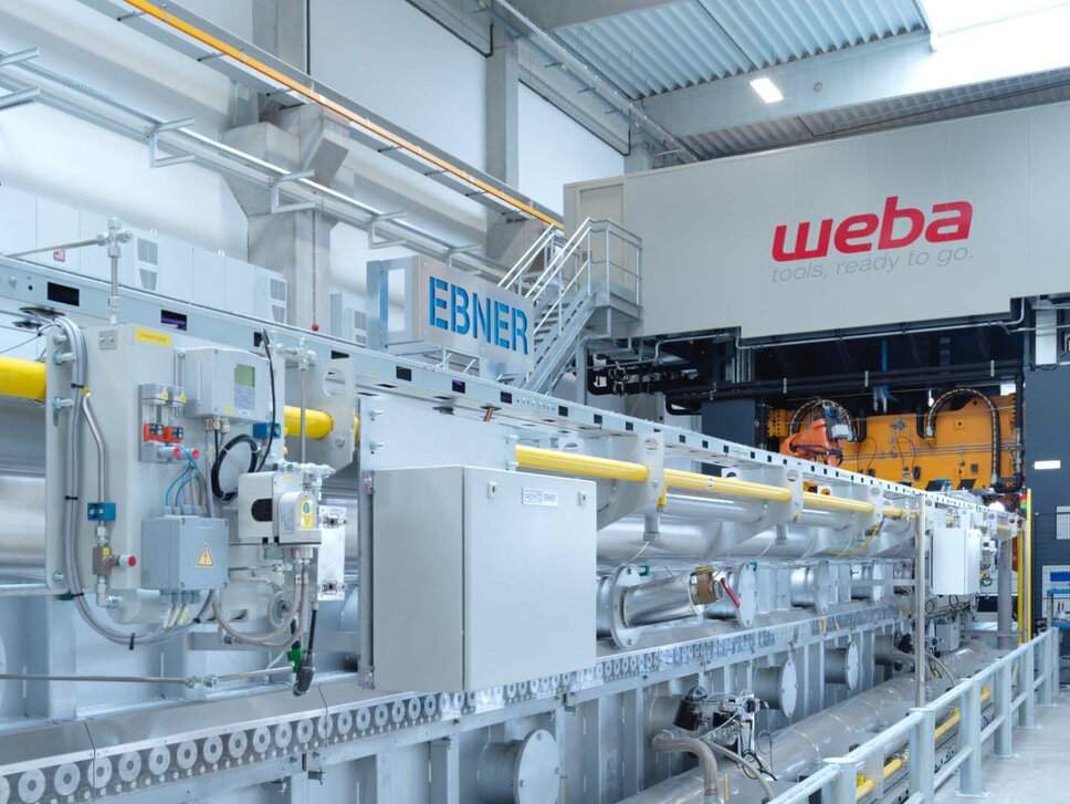 Foto einer Warmumformanlage von weba Werkzeugbau, auf dem ein 22 Meter langer Rollenherdofen und eine 1.600 Tonnen Umformpresse zu sehen sind. Die Anlage zeigt die industrielle Fertigungstechnik der Warmumformung in Aktion, wobei der Ofen für die Erwärmung von Metallplatten vor der Umformung verwendet wird und die Presse das erwärmte Metall in die gewünschte Form bringt.