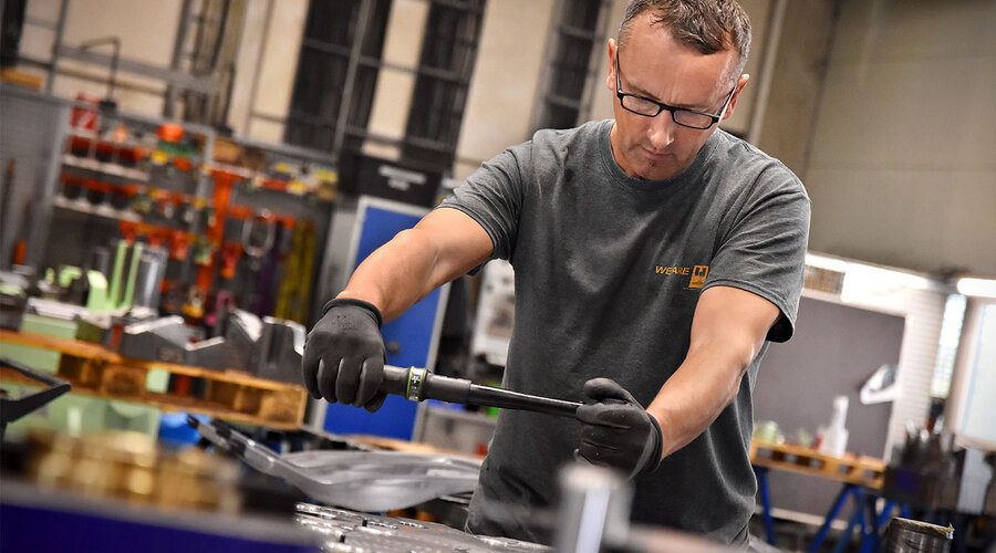 Das Bild zeigt einen Werkzeugbautechniker, der konzentriert an der Montage eines Stanzwerkzeugs arbeitet. Seine Hände sind geschickt damit beschäftigt, die verschiedenen Komponenten des Werkzeugs zusammenzufügen. Im Hintergrund sind Werkzeugteile und Montagewerkzeuge zu erkennen. Die Szene vermittelt ein Gefühl von Handwerkskunst und Präzision bei der Herstellung hochwertiger Werkzeuge.