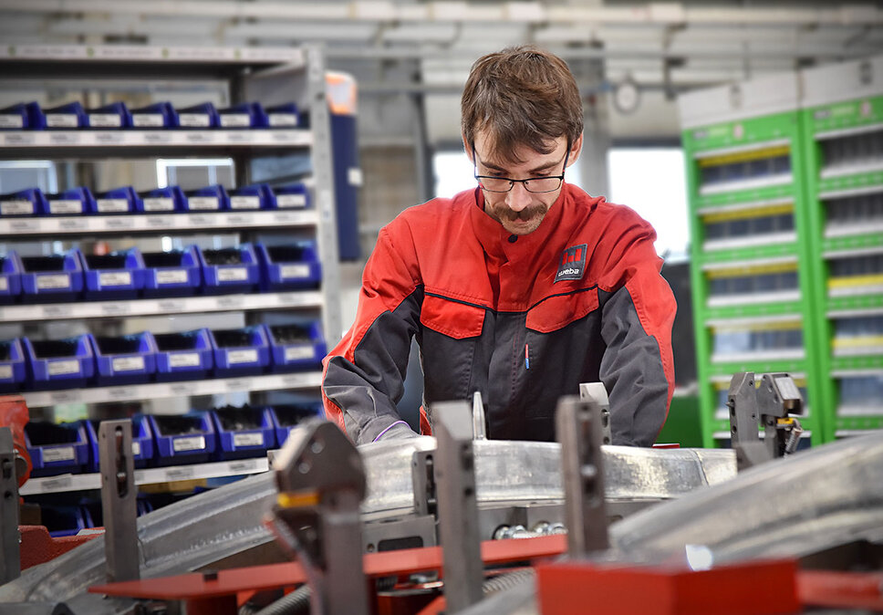 Ein Werkzeugbautechniker baut konzentriert ein Presswerkzeug für die Umformung von Strukturbauteilen zusammen; Im Hintergrund sind unscharf Regale mit Hilfs- und Betriebsstoffen 