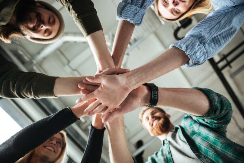 Ein junges Team lächelt und legt die Hände in einem Kreis übereinander. Das Bild wird symbolisch für Arbeitskultur bei weba und Berufseinstieg in Steyr verwendet.