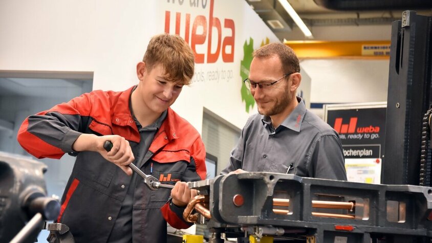 Ein Lehrling in der Werkzeugbautechnik arbeitet an einer Miniatur eines Umformwerkzeugs, während der Ausbilder aufmerksam zuschaut. weba Werkzeugbau bietet Lehre in Steyr in der Metalltechnik an.