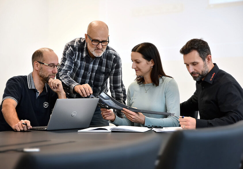 Four people are seated at a meeting table: one person is working on a laptop, another is attentively reviewing documents, while two others are engaged in a discussion about a stamping part. The scene depicts a focused and dedicated work environment. Experienced professionals can find their dream job at weba in Steyr.