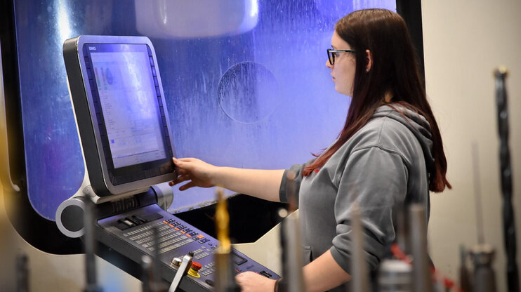 Eine Frau mit langen braunen Haaren und Brille arbeitet an einer CNC-Maschine. Sie trägt einen grauen Pullover und konzentriert sich auf das Bedienen des Touchscreens der Maschine. Im Hintergrund ist das Innere der Maschine zu sehen. Das Foto wird beim Stellenangeboten in Steyr als Symbolfoto für Zerspanungstechnik verwendet.