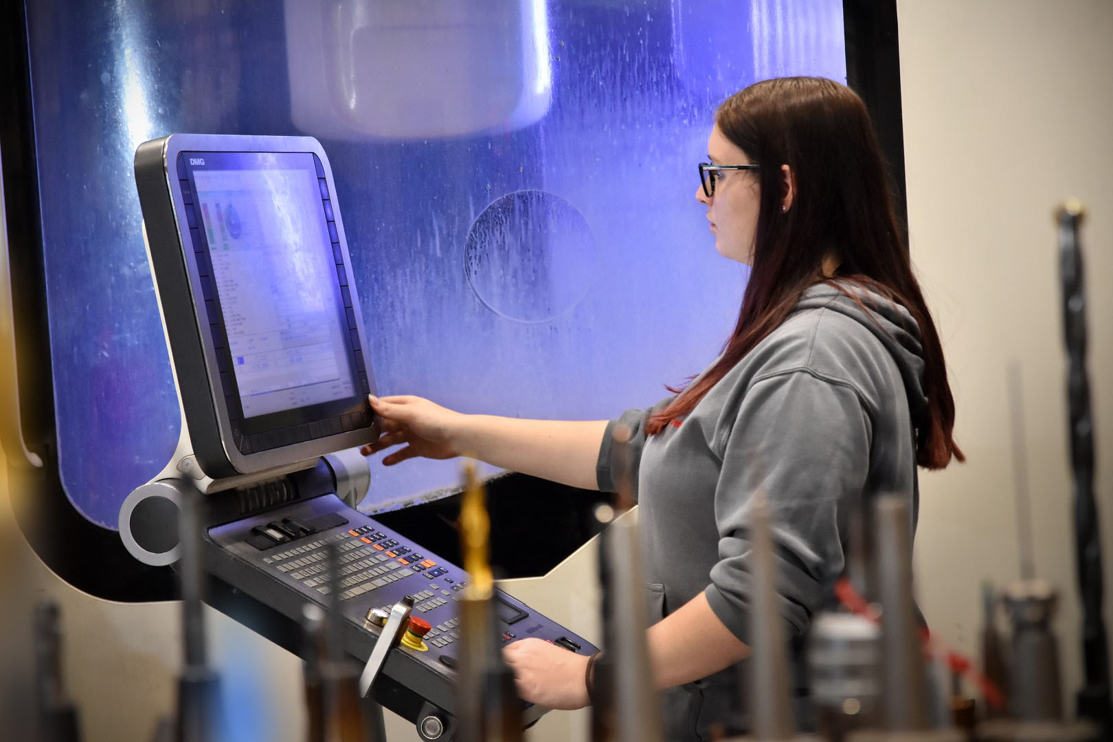 Eine Frau mit langen braunen Haaren und Brille arbeitet an einer CNC-Maschine. Sie trägt einen grauen Pullover und konzentriert sich auf das Bedienen des Touchscreens der Maschine. Im Hintergrund ist das Innere der Maschine zu sehen. Das Foto wird beim Stellenangeboten in Steyr als Symbolfoto für Zerspanungstechnik verwendet.