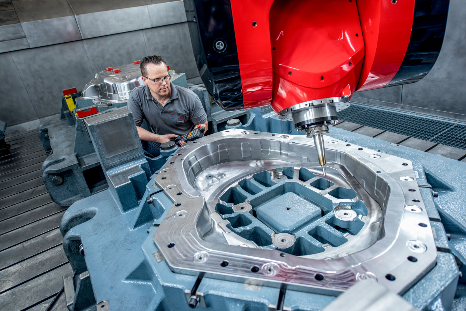 Ein Facharbeiter bedient eine hochmoderne CNC-Fräsmaschine mit einem großen Werkzeugbauteil. Die glänzende Metalloberfläche des Bauteils zeigt präzise Zerspanungsergebnisse, während die rote Fräseinheit prominent im Vordergrund zu sehen ist.